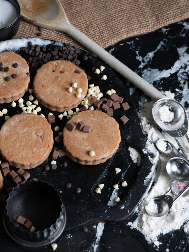 Triple Chocolate Welsh Cakes on a black griddle with loose flour and chocolate chips scattered around. 