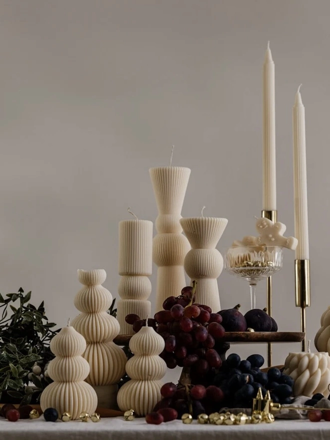 Signature Christmas Tree Sculpture Candle. A styled holiday table with cream sculptural candles, including ribbed trees, totem pillars, braided pillars and tall tapers in gold holders, alongside grapes, figs and greenery. The coordinating collection creates a sophisticated, warm display for the festive season.