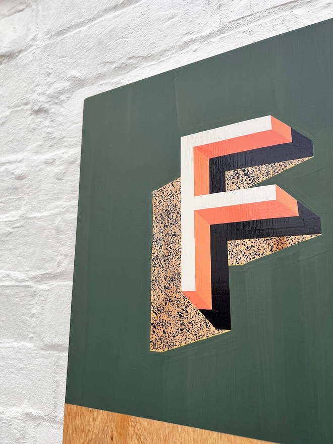 Close up of the letter F from FOOD hand painted sign in coral, green and aubergine, against a white brick wall, at an angle. 