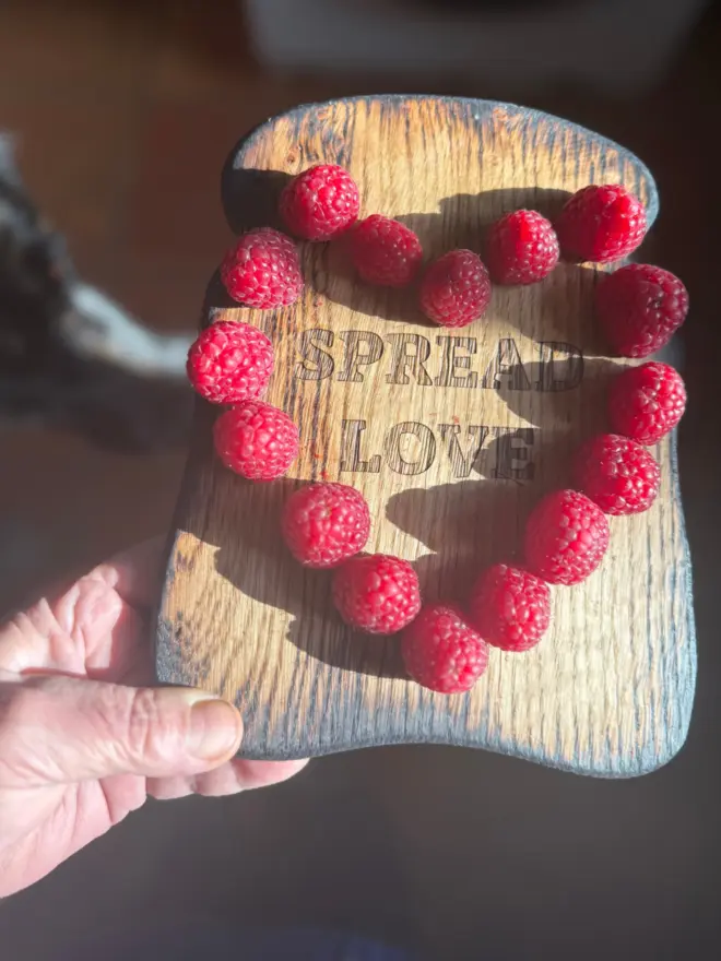 Small oak toast shaped serving board engraved with spread love surrounded by raspberries in a heart shape 