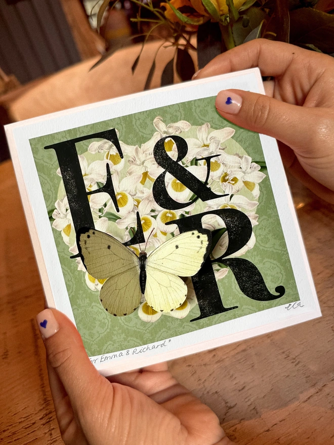 Hands holding a mint green wedding, monogram card, showing the initials E and R. The initials are black and placed on a white orchid heart. A white 3D hand cut paper butterfly sits on the bottom left of the heart. The card has the option to personalise further with the names of the couple, handwritten and the wedding date in the bottom, border of the card.