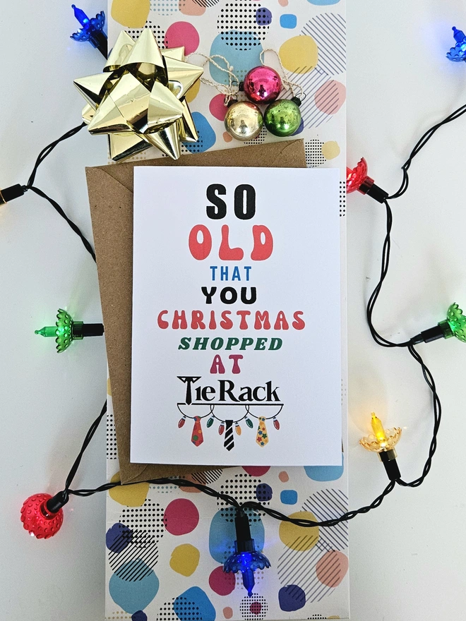 A photograph of the front of a humorous A6 greeting card illustrated using brightly coloured text and the retro shop logo. The text says so old you Christmas shopped at Tie Rack with a string of fairy lights and 3 colourful ties dangling from it