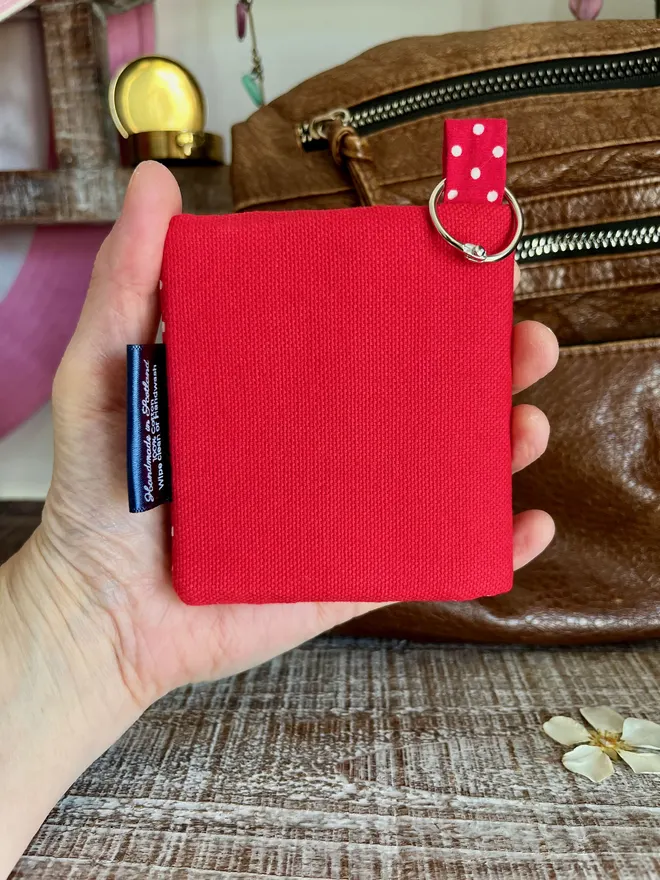 Small red card wallet held in the palm of a hand, showing a plain back and silver keyring attachment.