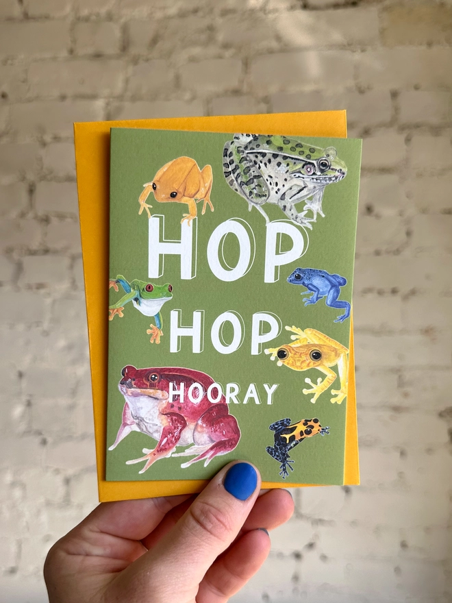 a green greetings card featuring a selection of frog illustrations with the text “hop hop hooray” with a deep yellow envelope