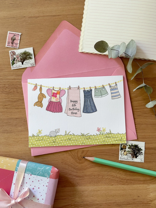 A personalised birthday greetings card with an illustrated washing line of children's clothes lays on a pink envelope on a wooden desk.