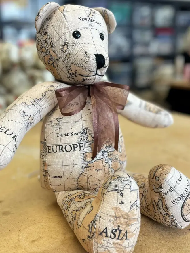 Teddy bear made from map-print fabric sitting upright on a wooden table, wearing a brown bow.