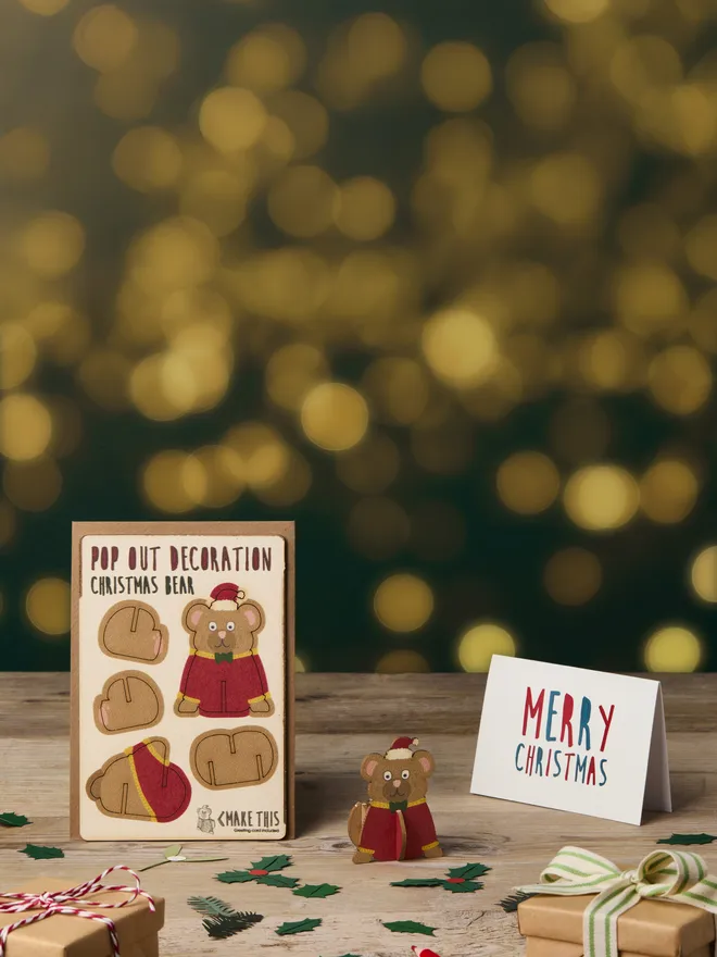 Pop out laser-cut bear Christmas decoration and Merry Christmas card and brown kraft envelope on top of a wooden desk in front of a festive lights background
