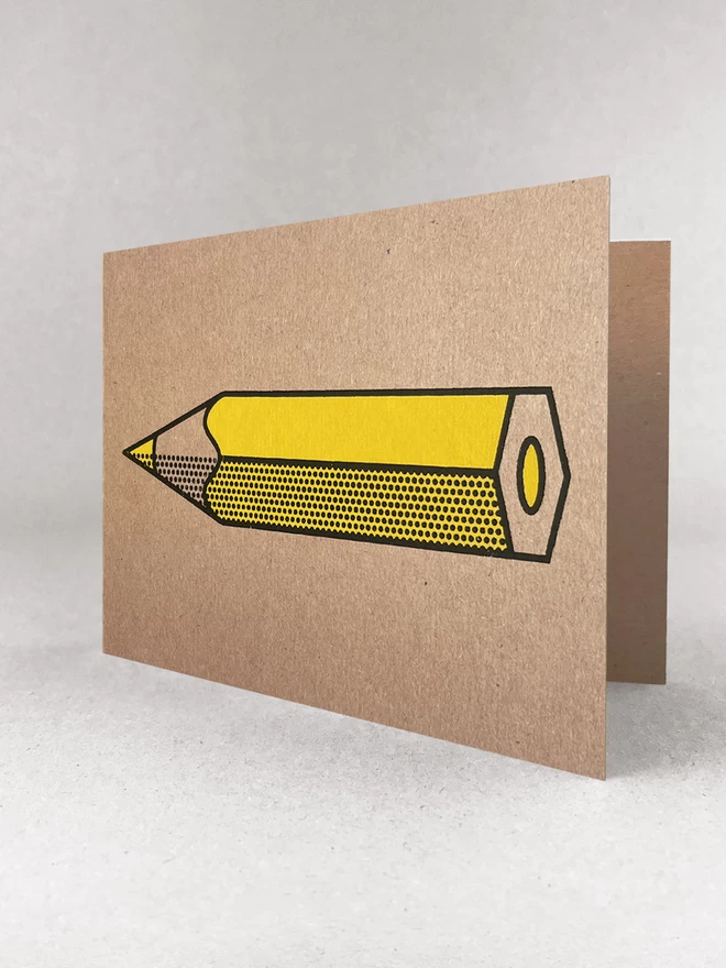Kraft brown card, stood in a light grey studio background. The design is a yellow pencil, horizontal with a black outline and halftone detail. The card is at a slight angle so we can see it open a bit.