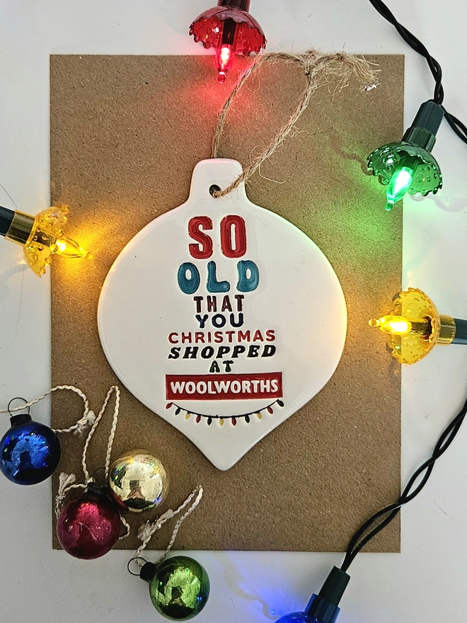 A handmade flat white ceramic bauble which measures 10cm by 8cm with So old that you Christmas shopped at Woolworths written on it in colourful glazes and retro fonts with a little string of colourful fairy lights stamped into the clay.