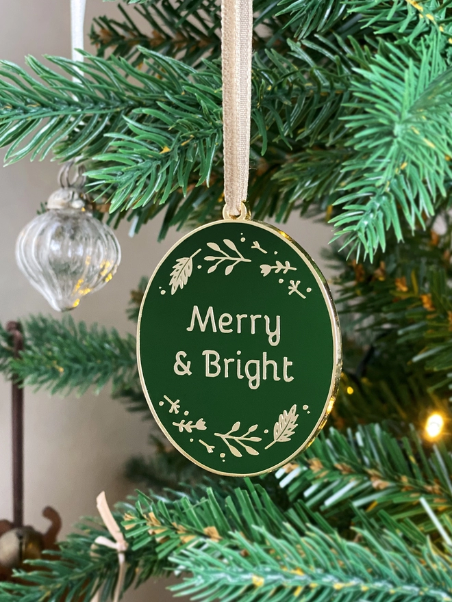A deep green and gold enamel Christmas decoration, with the words Merry & Bright surrounded by golden flowers, hangs on a Christmas tree.