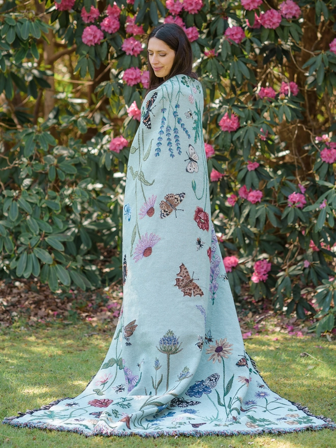 Woman standing with the 'Pollination Meadow' Recycled Cotton Blanket by Arcana draped over her shoulders.