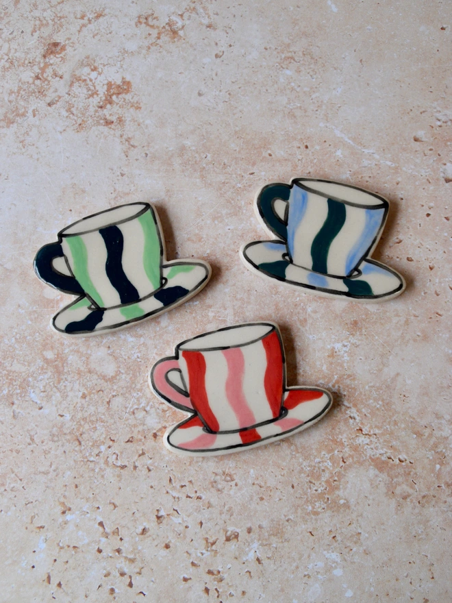 Handmade ceramic magnets shaped like cups and saucers, decorated with playful stripe patterns in red, pink, blue and green tones, arranged on a textured neutral background.