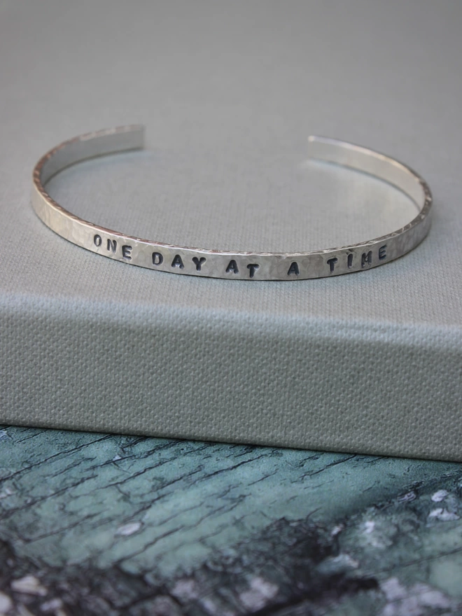 A single sterling silver hammered cuff bangle with the words 'one day at a time' resting on a green cardboard box lid on a green and white background.