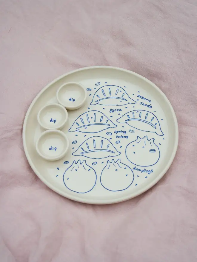 Dumplings Dippy Dinner Plate, a dipping plate sitting on a purple bed cloth. It is covered in blue outlines for dumplings. 