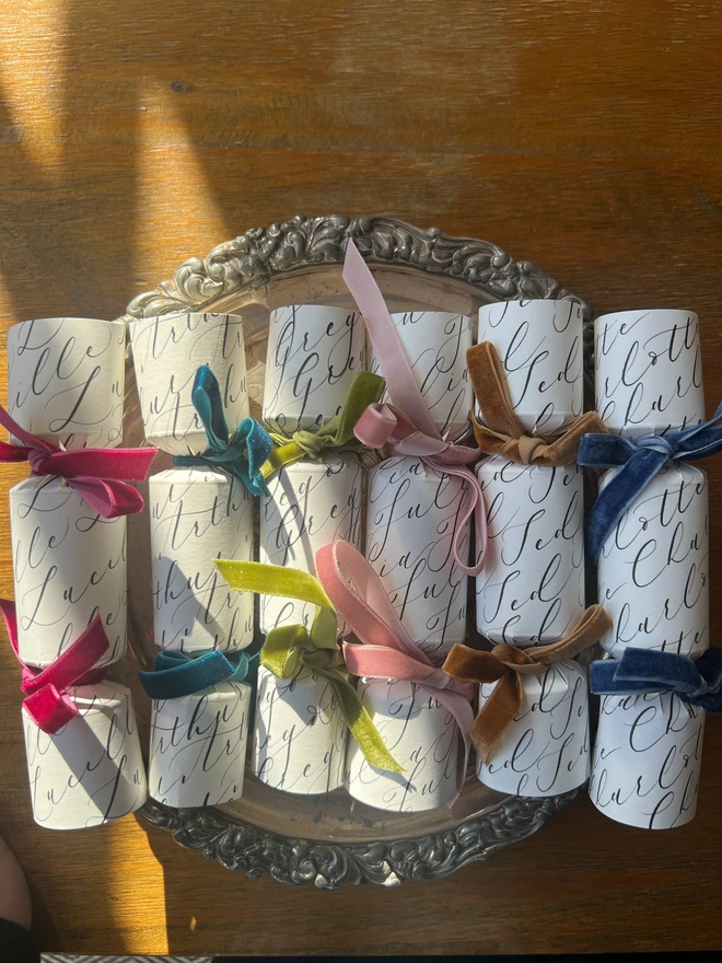 Selection of crackers personalised with calligraphy names tied with green, pink, mustard, blue, teal, rose, cerise velvet ribbon, laid on a silver tray on a wooden table