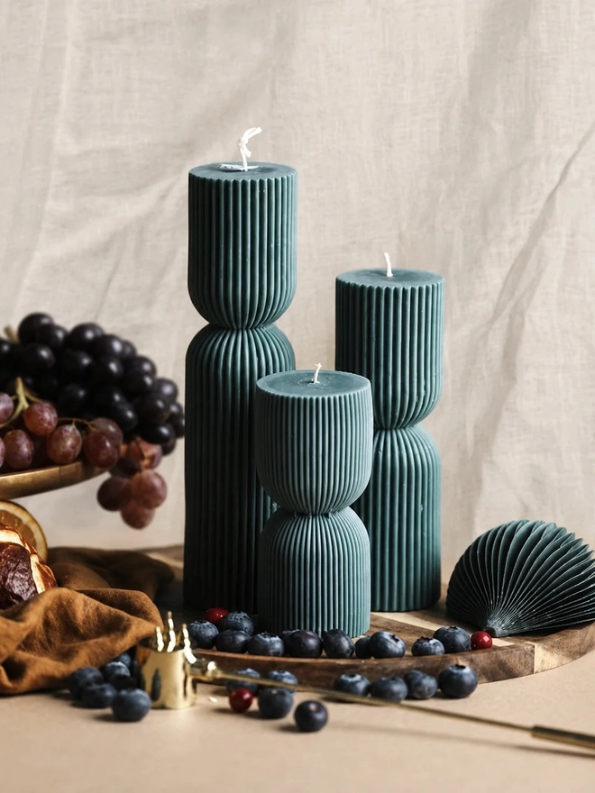 Forest Green Christmas Cracker Candles (Set Of 3). Close-up of three teal fluted pillar candles in graduating heights, styled with grapes, blueberries, cranberries and a brass snuffer against soft linen. The fine vertical ridges give these handmade candles an elegant, modern finish.