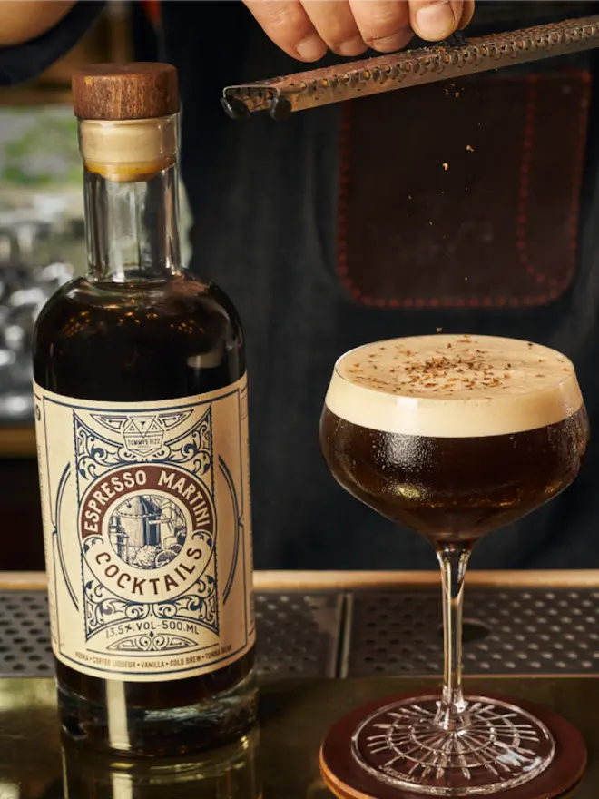 Tommy's Fizz Espresso Martini Cocktail, a bottle of Espresso Martini next to a filled coupe glass being garnished with coffee shavings.
