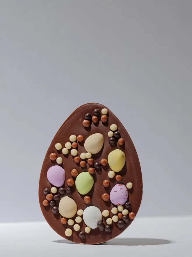 Large milk chocolate egg-shaped tablet decorated with pastel mini eggs and assorted chocolate pearls, standing upright on a plain white background.