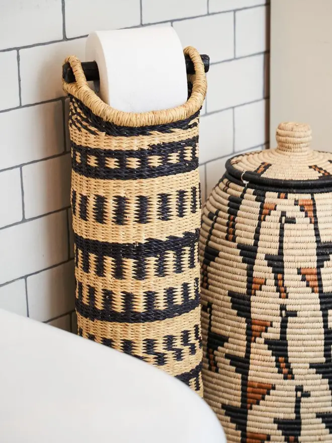 Ghanaian Hand Woven Tubular Toilet Roll Holder a toilet roll holder sitting in a bathroom. It sits in front of a plain backdrop accompanied by a basket. 