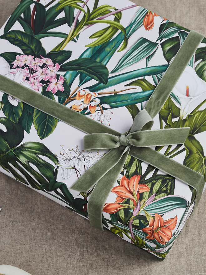palm house tropics floral gift wrap sheet. a box wrapped in white illustrated leafy gift wrap with a purple ribbon.