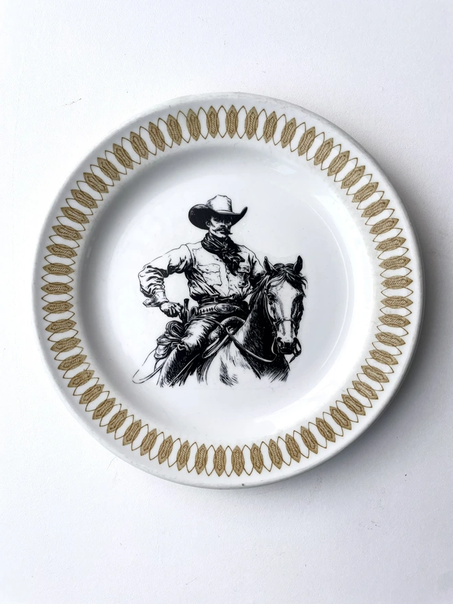 vintage plate hanging on a white wall, it has a decorative border of grey and blue leaves, with a printed vintage illustration of a cowboy in the middle 