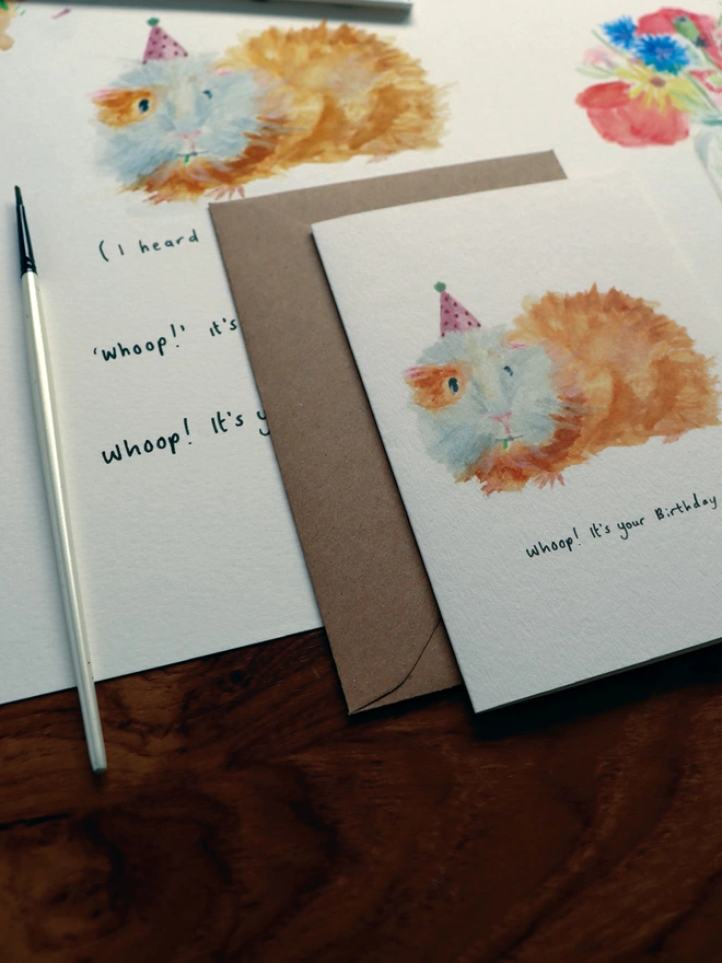  Close Up Slightly Out Of Focus Shot Of The Guinea Pig Greeting Card Sitting Along Side The Original Hand Painted Watercolour Illustration, Paintbrush and Palette