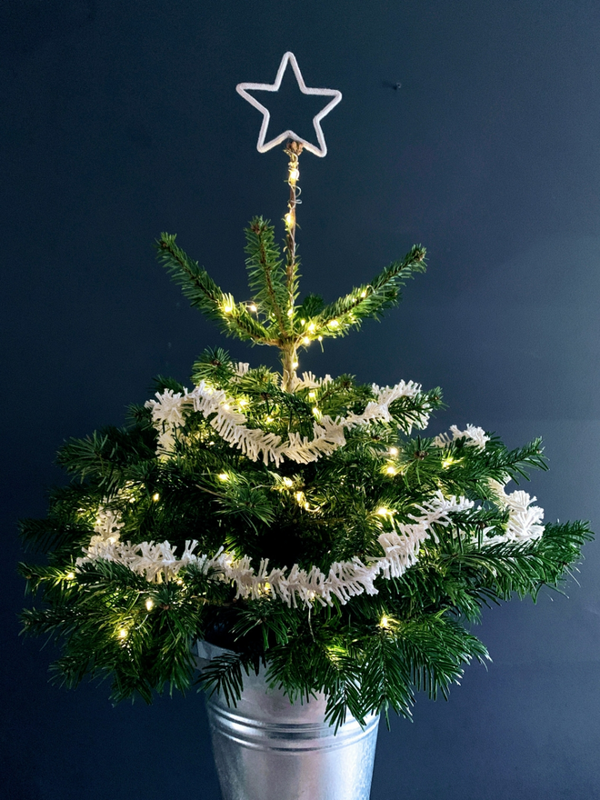 White Cotton Strinsel (plastic free string tinsel) on a star-topped green Christmas tree against grey wall