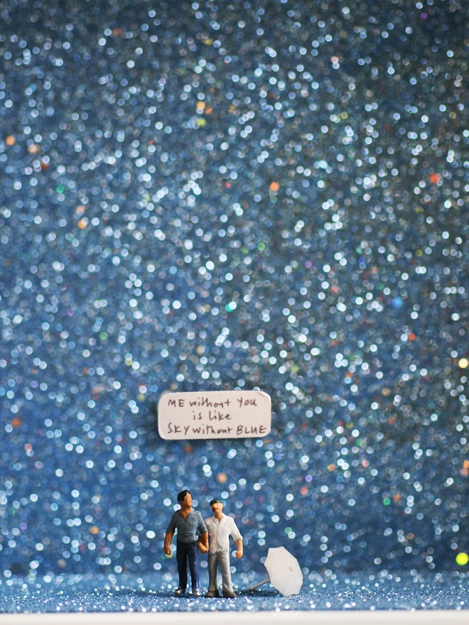 Miniature scene in an artbox showing two tiny men walking hand in hand, against a dramatic blue glittery backdrop evoking a rainstorm. The words “Me without you is like sky without blue” are written on the back wall