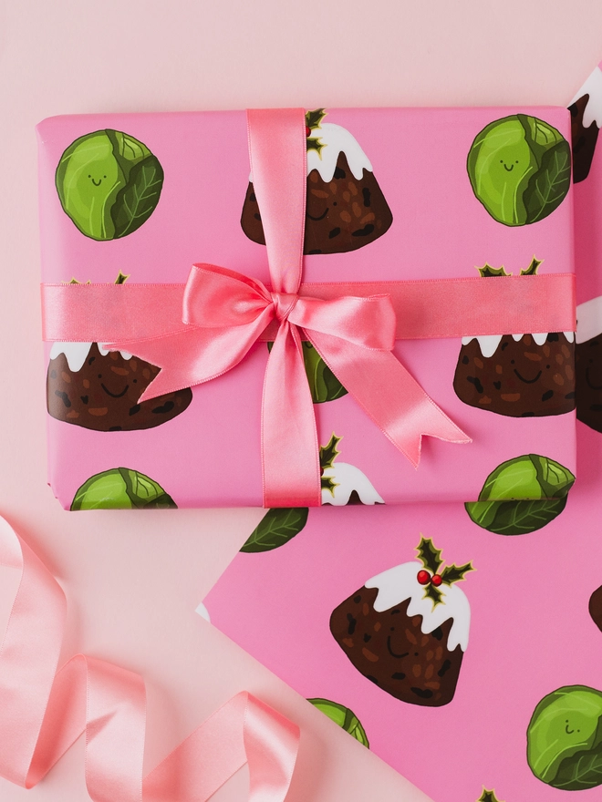 Pink Christmas wrapping paper featuring a repeating pattern of hand-drawn Christmas puddings with white icing and holly and smiling green Brussels sprouts.