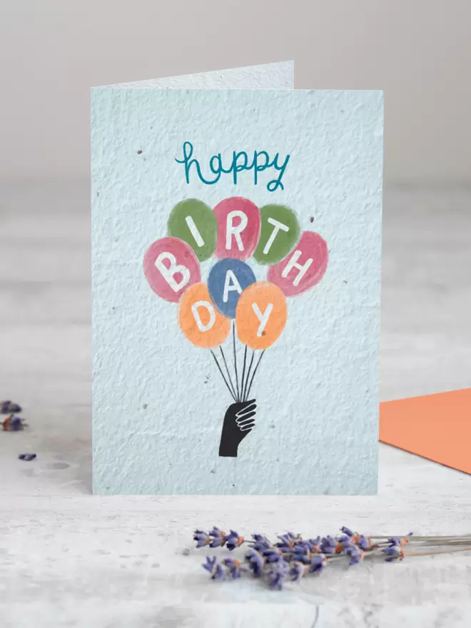Plantable Card with an an illustrated hand holding balloons with a letter in each balloon spelling out Happy birthday standing up with a sprig Lavender in the foreground