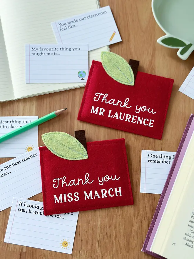 Two handmade red felt apple-shaped envelopes with green felt leaves and brown stems, each reading "Thank you MR LAURENCE" and "Thank you MISS MARCH", surrounded by illustrated prompt cards with child-friendly questions, displayed on a wooden desk with a pencil, notebook, and book. It's a charming personalised, handmade teacher thank you gift.