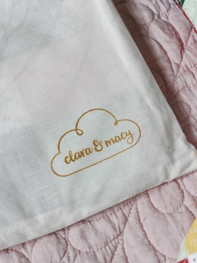 A cotton dustbag with the Clara and Macy logo in one corner rests on a pink quilt.