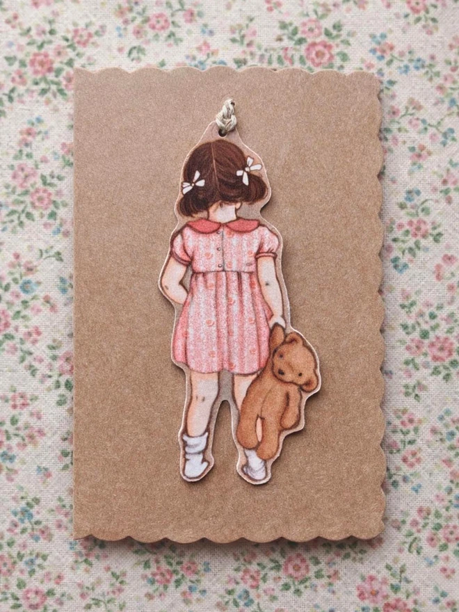 a scalloped kraft greeting card featuring a die cut swing tag of a little girl with bunched drawn from behind with a teddy