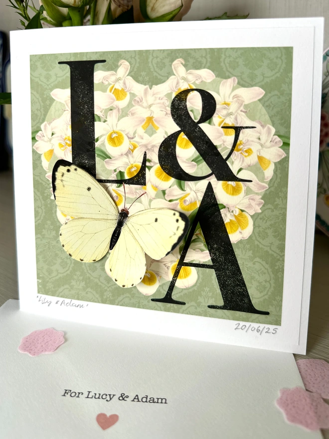 A mint green wedding, monogram card, showing the initials L and A. The initials are black and placed on a white orchid heart. A white 3D hand cut paper butterfly sits on the bottom left of the heart. The card has the option to personalise further with the names of the couple, handwritten and the wedding date in the bottom, border of the card. The envelope included, has the couples names printed.
