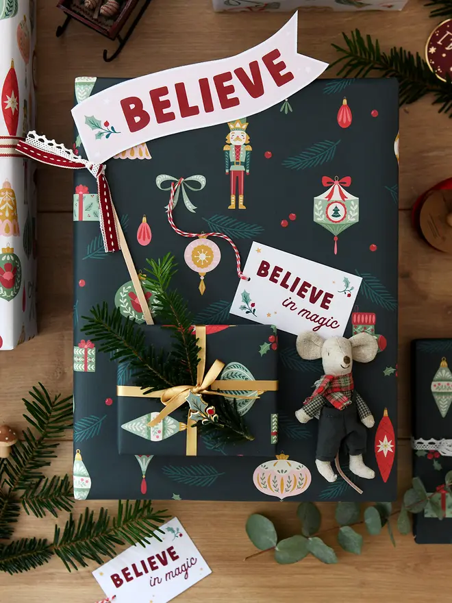 A stack of Christmas gifts wrapped in nutcracker wrapping paper are on a wooden floor. A Believe flag is tucked into the ribbon, and a small mouse toy lays beside it.