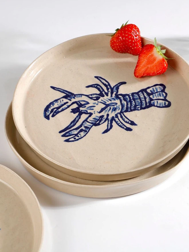Handmade Ceramic Side Plate With Blue Lobster Design, a side plate with a blue lobster design sitting on a white table surface accompanied by a raspberry. 