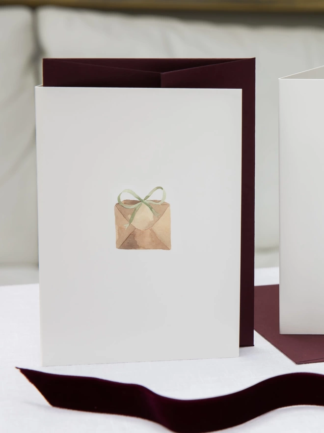 Greetings card with a watercolour illustration of a brown paper package tied up with a green bow which comes with a burgundy grape envelope