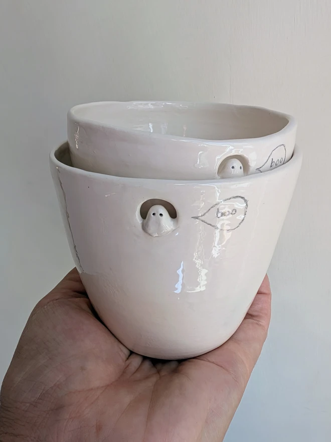 Small and large ghost cups with the smaller inside the larger, in white handmade ceramic with a tiny sculpted ghost in a cut out near the top of the beakers with a hand painted bubble containing the word boo, pottery tumbler for halloween