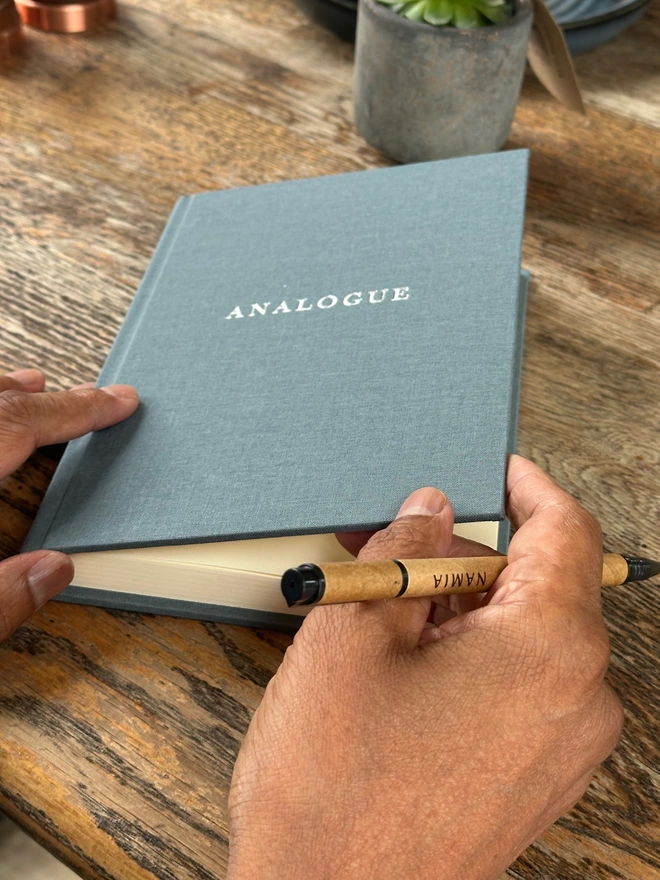 A notebook covered in grey linen and printed with 'Analogue' in gold lettering is held in a man's hands, along with a pen.