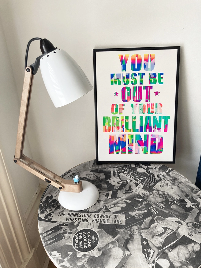 Framed multicoloured typographic poster with song lyric by the band, Furniture - "You must be out of your brilliant mind”.  The print sits on a round table decoupaged with 1970's wrestlers, next to an anglepoise lamp.