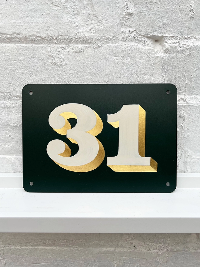 Hand painted house number against a white brick wall, number 31 in serif style in off white and directionally burnished 23 carat gold leaf, on anthracite grey background. 