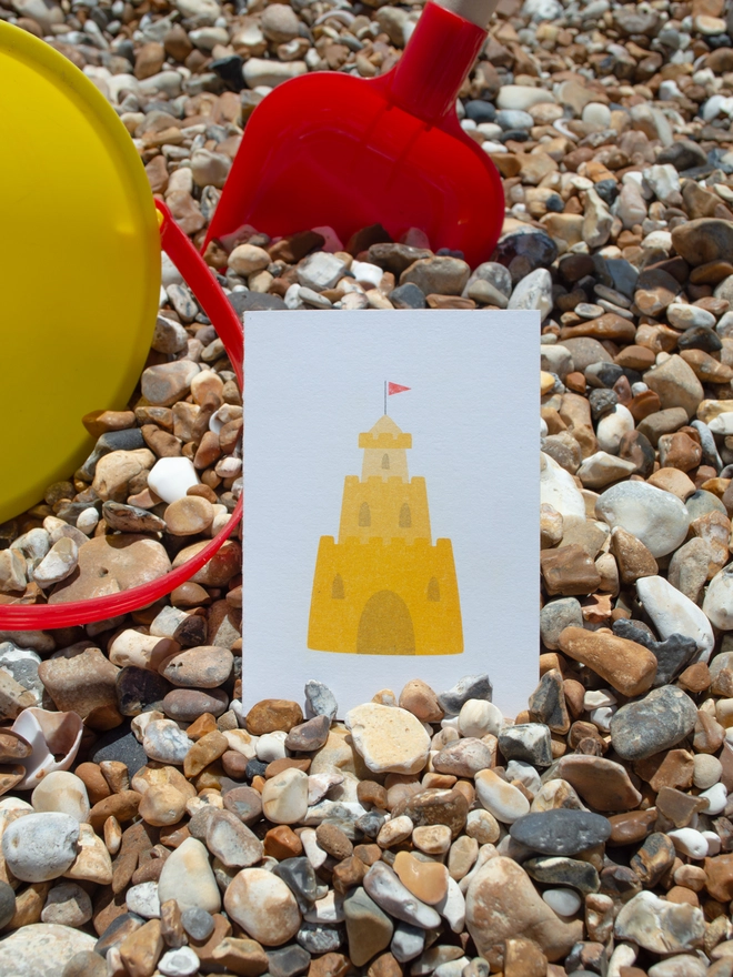 A pebbly beach is shown in the background with a bucket and spade positioned next to a small white greeting card which has a 3 tier yellow sandcastle printed onto it with a small red flag on the very top tier.