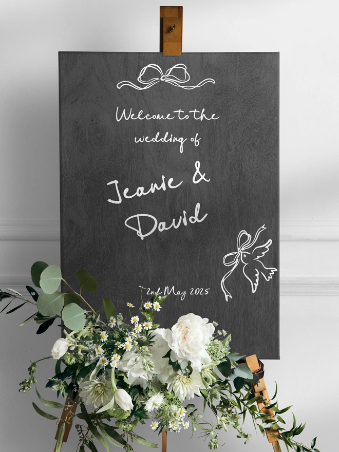 Black board on a wooden stand with the words Welcome to the wedding of Jeanie and David and the date of the wedding in white lettering, at the front of the board is a boquet of white flowers and green foliage