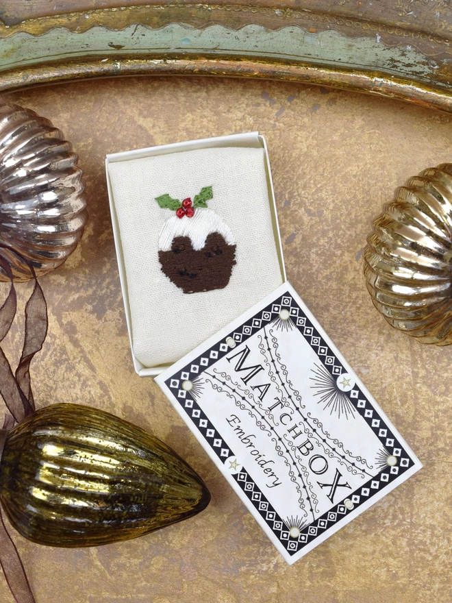 A Classic Christmas Pudding embroidery, inside a Matchbox displayed on a gold background with glass baubles.