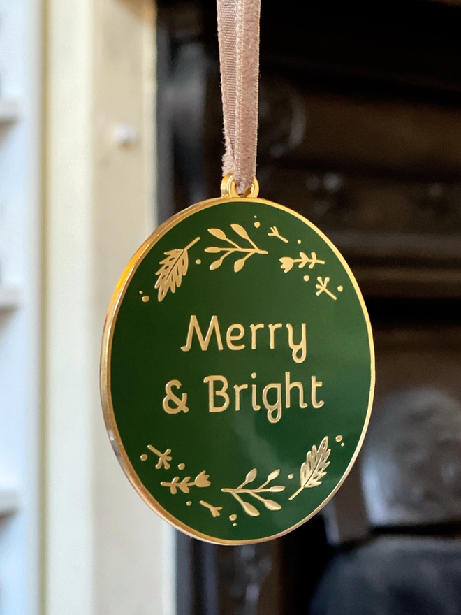 A deep green and gold enamel Christmas decoration, with the words Merry & Bright surrounded by golden flowers, hangs in front of a fireplace.