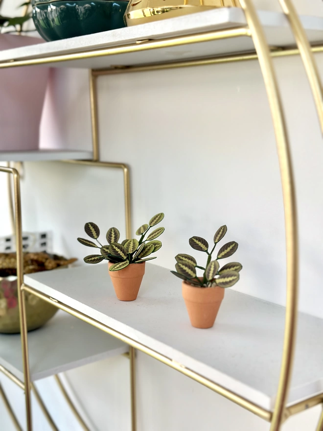 2 miniature replica Maranta Prayer Plant paper plant ornaments in terracotta pots sat on a shelf of a gold circle shelving unit with pther pink and gold ornaments in the background