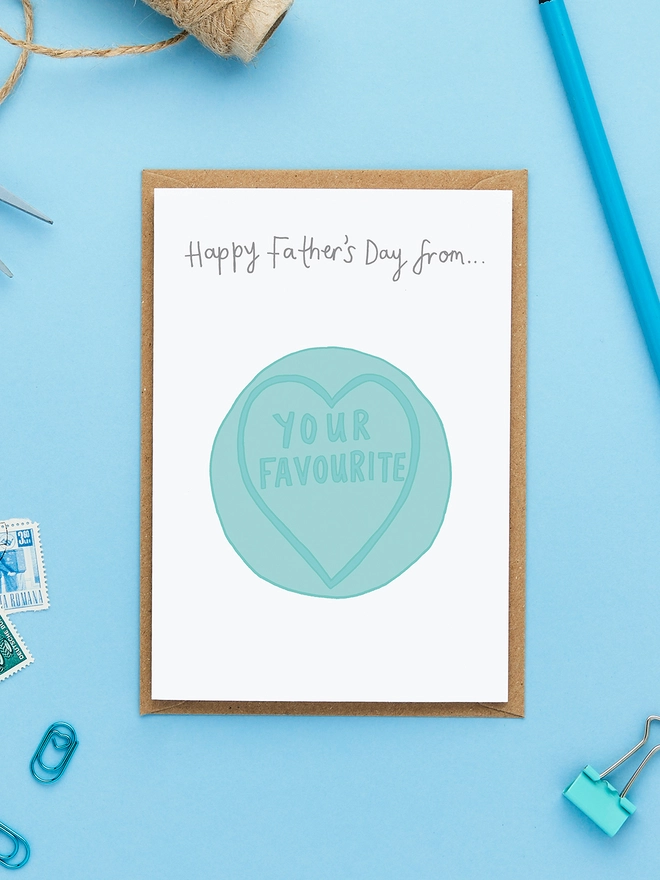 Greeting card styled on blue background with twine, pencil and stamps, featuring a blue love heart sweet that says "Your Favourite" and the caption "Happy Father’s Day from…".