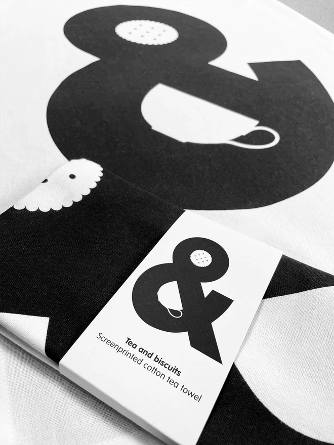 A close up of a folded tea towel lies on an opened out tea towel - the design is a giant black ampersand which has a cup and a biscuit hidden in the negative spaces. The folded tea towel has a belly band also showing the complete design.