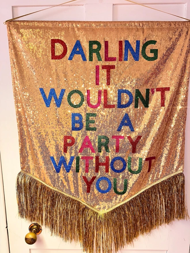 A maxi customisable banner hangs from a hook. It has a gold sequin background and a gold tinsel trim along the bottom. The text is multicoloured and says 'DARLING IT WOULDN'T BE A PARTY WITHOUT YOU'