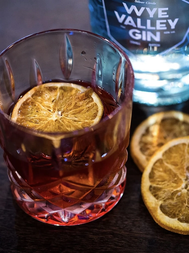 Damson Negroni, in a glass next to a bottle of spirits, with a slice of orange. 